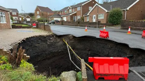 Cables can be seen in the deep sinkhole, with levels of soil visible inside the hole. Paving from the bungalow driveway hangs over the edge on one side and sections of tarmac from the road also hang over the hole. There are safety barriers in place. A number of other homes can be seen on the opposite side of the road. 