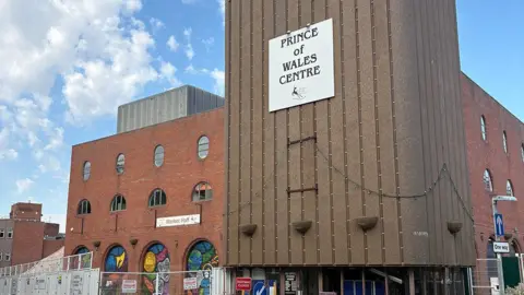 A large red brick building with multiple sections set back from one another and a ramp to the right side. There is a sign above the glass entrance doors which reads Prince of Wales Centre. The section to the left of the entrance has a number of archways containing colourful murals.
