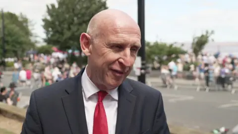 Healey wears a red tie, white shirt and dark jacket. He squints in the sun. Crowds can be seen in the background.
