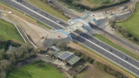 National Highways A bird's eye view is seen of the A30 Chiverton to Carland Cross road and bridge, surrounded by fields and with cars on it the road.
