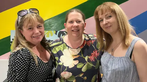 A blonde woman in a black and white dotted shirt, is standing next to a blonde woman in a flowery print shirt. On her right is another blonde woman wearing a striped white and dark navy top. 
