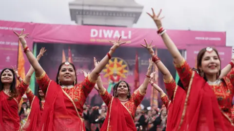 PA Media Four women standing in a dance formation and dressed in red and gold trimmed saris and wearing golden jewellery on their heads raise their hands in the air.  A stage behind them is decorated with purple banners: one says 'Diwali'. In the background is the plinth of Nelson's Column. 