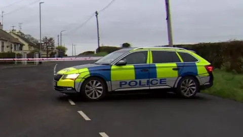 A police Car sits infront of a group of white brick houses. Inbetween the car and the houses is red and white police tape.