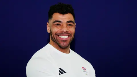 PA Studio shot of Taylor Lawrence in a white Team GB jersey. He has short black hair and is smiling for the camera.