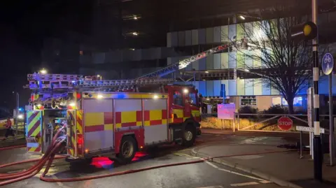 Matthew Webb/BBC A fire engine with hoses running from the back is parked facing a car park building with a ladder extended towards it from its roof.