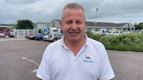 A man, with short grey hair, wearing a white t-shirt. He is standing in a carpark with a Lidl store behind him.