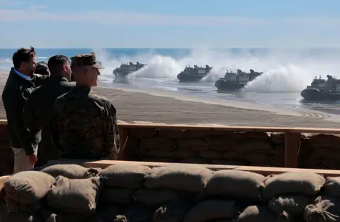 Getty Images JD Vance watches along side military members as boats reach the shores of a beach