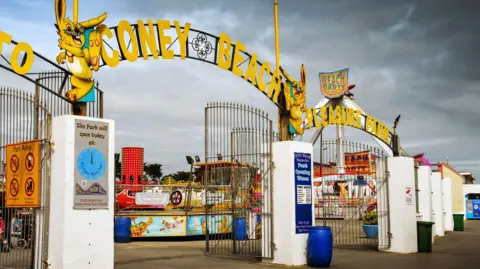 Entrance gates to amusement park with Coney Beach Pleasure Park in yellow letters on top of the gates and attractions visible behind 