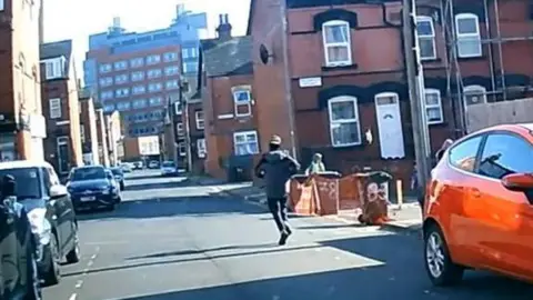 A suspect, dressed in a grey jacket and black trousers, runs down a residential street.