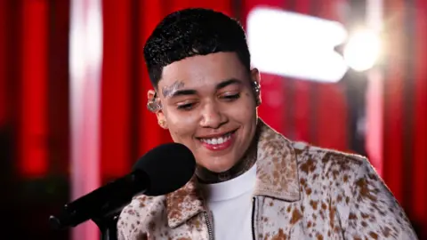KWN smiles as she performs in session for BBC Radio 1 Xtra in January 2026. She is wearing a dappled tan and brown jacket, and has in-ear monitors looped into her ears.