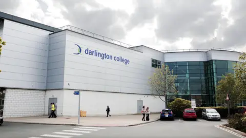 LDRS The outside of the Darlington College building, with the college's branding on the building. There is a grey sky above, and a few people walking around and some parked cars in the foreground. 