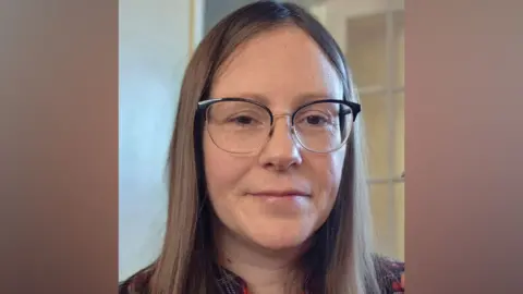 Lauren Rayner A woman with long brown hair smiles at the camera. She is wearing glasses and a red chequered top. 