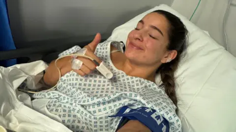 Betty Mukherjee A woman lying in a hospital bed wearing a patterned gown, with a blood‑pressure cuff on one arm and a pulse‑oximeter on a finger, giving a thumbs‑up amid nearby medical equipment.