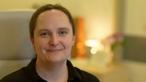 Nicola Dolden is smiling at the camera, with dark hair tied back, and is wearing a black top. The background is blurred out.