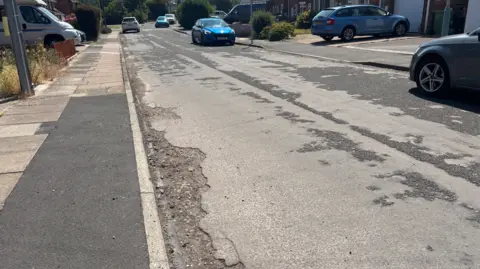 The photos shows the street which is considerably deteriorated. There's patches of black tarmac but most of it is grey. Next to the pavement the grey tarmac goes completely with stone and brick showing underneath. The pavement is also in a bad shape, patched up with tarmac in places with cracked slabs further down the street. Cars are on the road and the driveways of the homes.