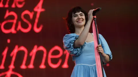 Jim Dyson/Getty Images A female singer wearing a blue dress smiling while holding a microphone and a microphone stand. 