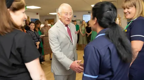 King Charles III dressed in a light grey suit shakes hands with hospital staff on a ward.