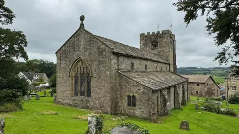 External view of St Wilfrid's Church which is in the village of Melling near Lancaster. Gravestones can be seen in the churchyard with fields in the background.