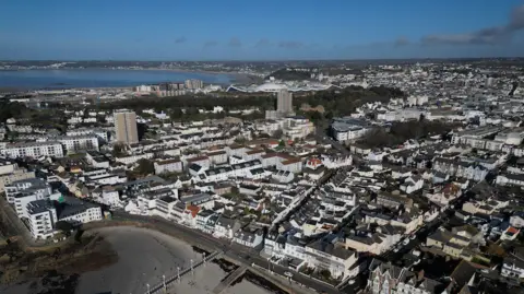 An aerial view of the south of Jersey - rows of houses and a beach in the foreground, sea can also be seen further afield. Blue skies.