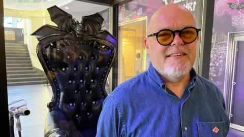 Jez Collins standing beside ‘The Throne of Ozzy Osbourne’, an ornate black chair with bat-wing details and skull armrests displayed in a glass case inside a museum exhibition space.
