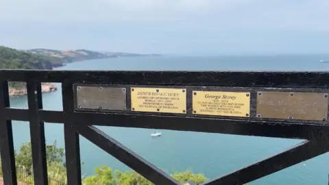 A row of commemorative plaques attached to a railing at Babbacombe Downs looking out to sea.