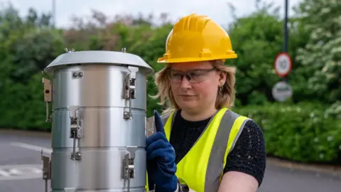 University of Southampton Dr Nat Easton wears a high-vis, yellow jacket and a safety helmet as she attends to a conical, metallic chamber beside a road.