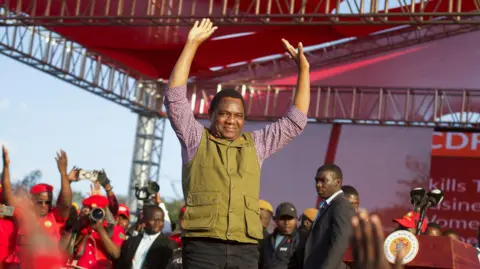 Zambia's President Hakainde Hichilema in green half-coat and black trousers lifts his hands to greet United Party for National Development supporters