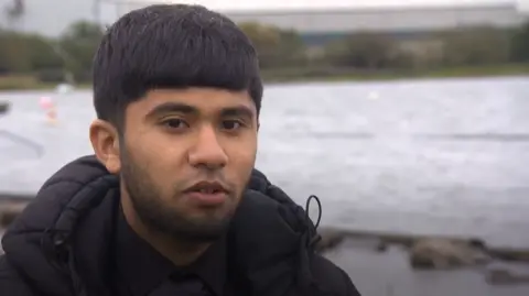A young Asian man, with black hair and a light black beard, speaks to the camera standing in front of a body of water. He is wearing a black winter coat over a black shirt.