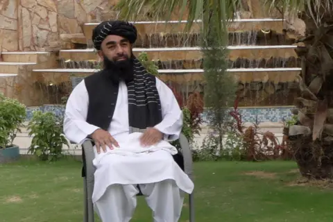 A man in traditional white Afghan tunic and trousers with a black waistcoat and headdress sits in a chair on a lawn. Behind him are a large fountain, neatly laid stone steps, shrubs and a tree.
