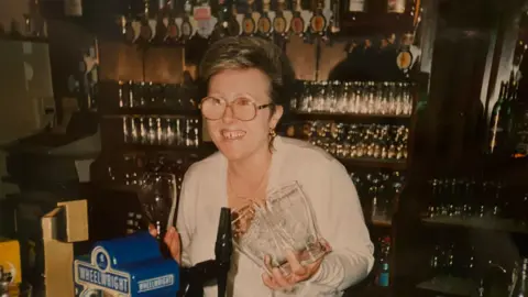 Linda Oliver A woman smiling, holding pint glasses stands behind a bar. this is Linda but she's younger, has brown hair and bigger glasses. The photograph is older.