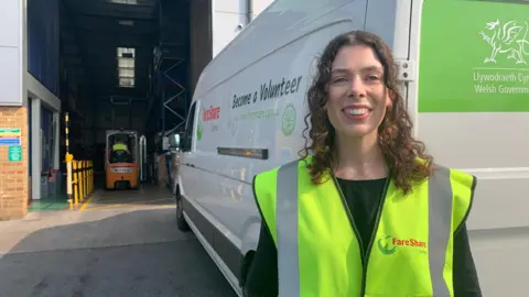 Katie in front of white delivery van by a warehouse in yellow high vis jacket. She has long, dark, curly hair and a forklift truck is visible behind.