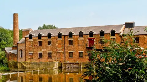 Picture of Thwait Mill in Leeds. It's a former mill building, which is bricked and has multiple windows on it. A chimney to the left. The mill sits on the banks of the River Aire, which is pictured.