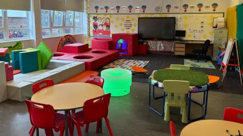 A brightly decorated classroom with round tables and red chairs in the foreground, soft play equipment and padded climbing blocks along the left wall, a glowing green sensory light table in the centre, and a large interactive screen with desks and colourful displays on the far wall. Hot‑air‑balloon artwork and children’s drawings are displayed above the screen, and the room features cushions, rugs, and various learning stations.