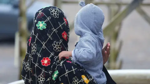 A woman wearing a head scarf with flowers on it holds a young child who has a blue and white stripey hooded jumper on.