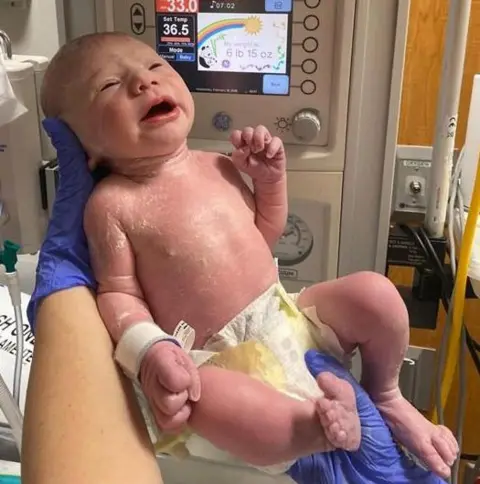 Maisie Savage Newborn baby in the foreground, wearing only a disposable nappy and supported by gloved hands in front of a neonatal warming unit. A hospital identification band is attached to the baby’s wrist. The warming unit’s display screen shows temperature settings and a weight reading, with control buttons and gauges visible around it. Medical equipment, tubing and wall-mounted hospital fittings appear in the background. The image is taken in a clinical hospital setting.