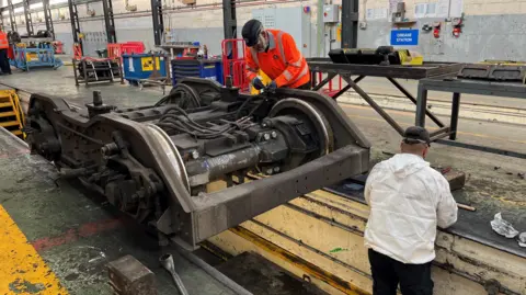 Two engineers working on a train bogey. The photo shows one man wearing a white jacket lower down on the train tracks and on the platform another man wearing a fluorescent jacket is working on the top section of the train bogey. In the background are metal work tables placed alongside each other.