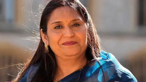 Seema Misra poses for a photo after receiving an OBE. She is wearing blue and has gold earrings and she is standing outside in the sunshine.