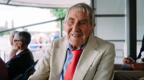 Tony has grey/white hair. He sits in the stands at the regatta wearing a white blazer, blue shirt, and red tie.