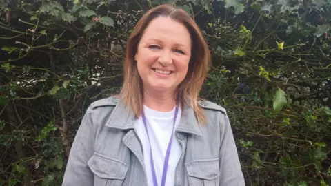 Lisa is standing outside in front of a bush. She has shoulder-length brown hair and is wearing a white top and a khaki jacket. She is smiling at the camera.