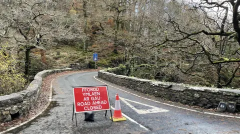 A red sign on a bridge that says "road ahead closed" and says the same phrase in Welsh. The road is narrow and has a deep right hand bend. There are trees surrounding the road on both sides, with a visible drop from the roadside on the right hand side.