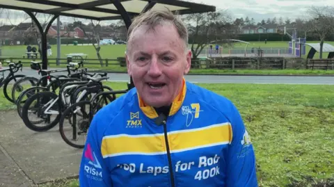 A man with brown hair stands on a grass verge surrounded by a bicycles. He is wearing a blue cycling jersey with a yellow and white stripe across the centre.