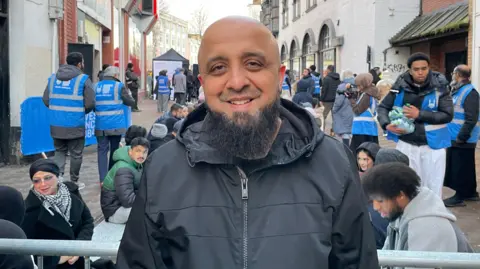 BBC Mohamed wearing a black jacket standing in front of a railing with people kneeling behind him