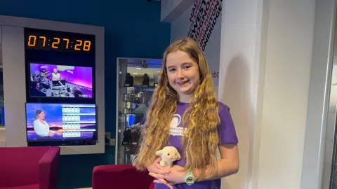 BBC The image shows a girl standing indoors in a radio studio environment. She has very long, wavy, light‑brown hair that extends well past the shoulders. They are wearing a purple T‑shirt featuring a white graphic and text on the front, along with black Nike trousers. On their wrist is a bright green watch. They are holding a small plush toy that looks like a pale-coloured rabbit.