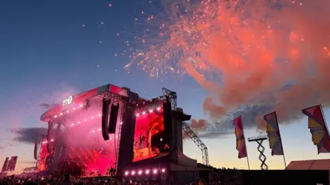 Olivia Dean is seen on stage on a screen, with pink lights and smoke in the sunset.