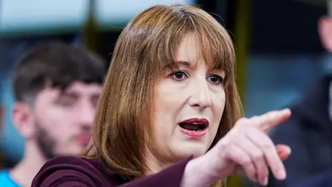 PA Media Rachel Reeves pointing into the distance, wearing a purple top with a man out of focus in the background.