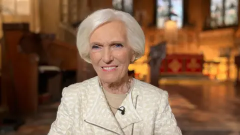 BBC Mary Berry smiles in a large church, which is blurred in the background behind her. She is wearing a tailored white and cream jacket and short white hair. 
