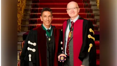 NHS Humber Health Partnership Professor Sunil Bhandari standing with Professor Mark Strachan. Professor Sunil Bhandari is wearing a black and red robe, holding a small gold medal which sits in a box. He has brown hair and is smiling at the camera. Professor Mark Strachan is wearing a black and red robe over a grey suit blazer, a white shirt and a red tie. He is bald and is wearing glasses. He is smiling at the camera. They are stood in front of a set of red-carpeted stairs.