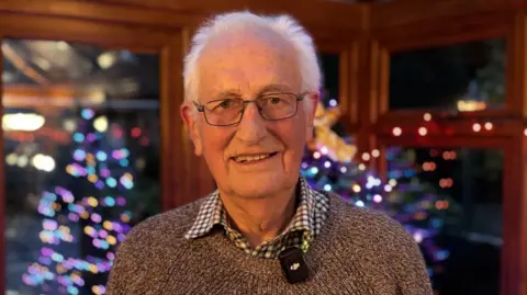 BBC Gerald is standing close to the camera wearing a brown knitted glasses and black framed glasses. Behind him, out of focus, is a Christmas tree reflected in a conservatory window.