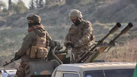 Reuters Two Syrian security force soldiers sit on top of a pick up truck armed with a  twin-barrelled heavy machine gun. Both men have masks on their faces and are dressed in full military fighting equipment and camoflage uniforms.