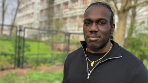 A man in a black top with a black three-quarter zip and wearing a gold necklace stands in front of a block of flats which are blurred in the background. 
